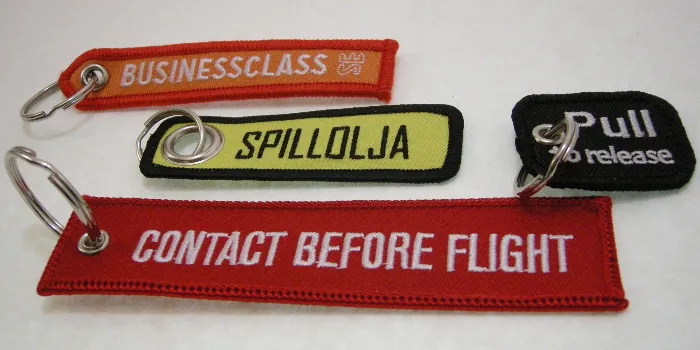Broderade nyckelringar med langetterad kant i olika versioner av den klassiska 'Remove Before Flight' taggen.