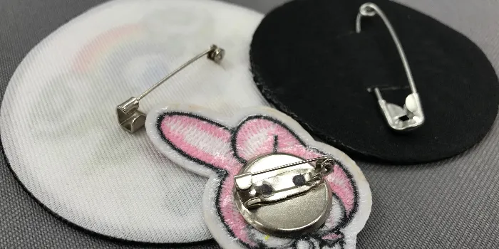 Baksidorna på tre broderade tygmärken med broschnål.
