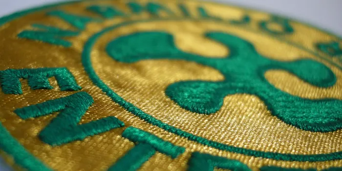 Ett brodyrmärke med bottenmaterial i guld metallic tyg och grön broderad text och logga.