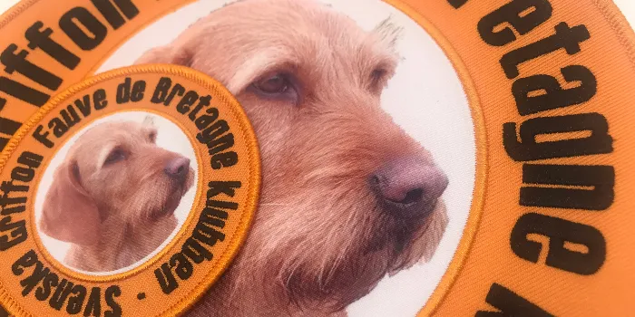 Stor och liten version av samma hybridtygmärke med fotografiskt tryckt motiv av en hund och svart broderad text runt omkring längs orange kant.
