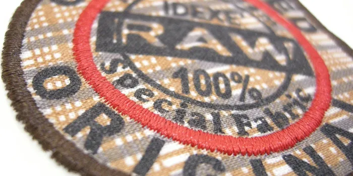 Hybridtygmärke med vävd rutig bakgrund och tryckt text i svart samt mörkbrun broderad kant och röd inre ring.