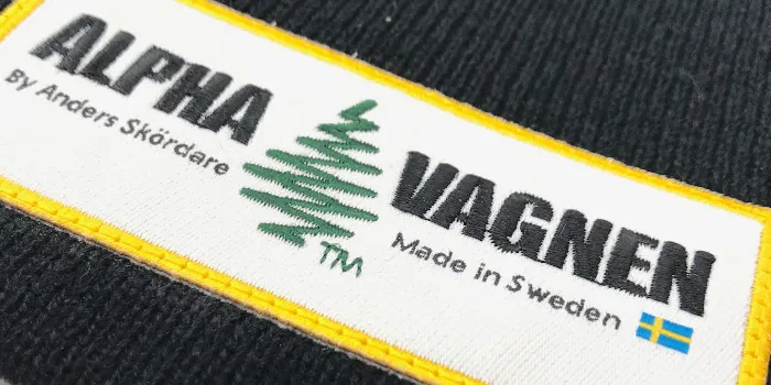 Hybridtygmärke där vi blandat broderad stor text och symbol med mindre text som vävts.