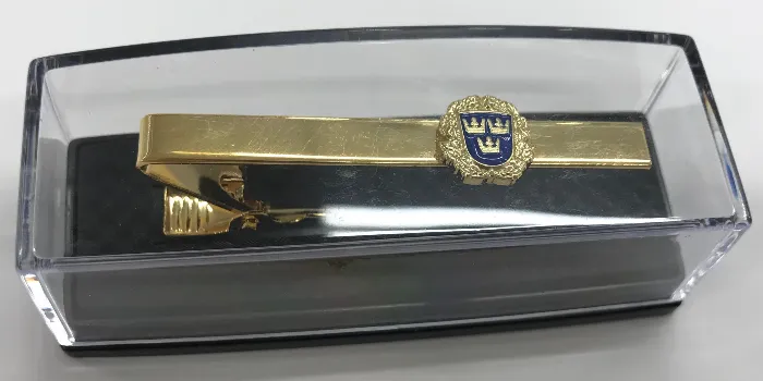 Slipsnål i guld försett med emblem med blå emaljfärg inuti ett genomskinligt etui.