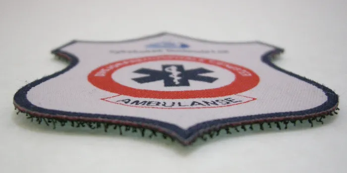 Sköldformat vävt tygmärke med kardborren på baksidan delvis synlig.