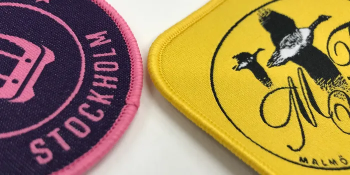 Närbild på två vävda tygmärken med 3 mm bred langetterad kant varav en i rosa kontrastfärg och den andra gul ton i ton.